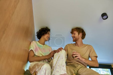happy asian woman and redhead man holding cups of coffee and sitting together at cozy bunk bed