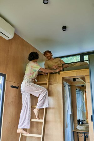 Photo for Happy redhead man looking at brunette girlfriend climbing on ladder of bunk bed, weekend getaway - Royalty Free Image