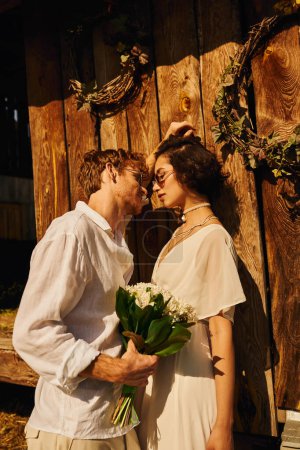 redhead man in sunglasses holding floral bouquet near asian woman in wedding dress near wooden barn