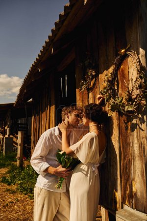 redhead man in sunglasses holding bouquet near pretty asian woman in wedding dress near wooden barn