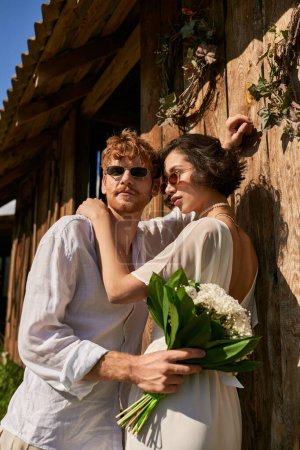 Photo for Asian woman in wedding dress hugging with groom in sunglasses near wooden barn, rustic wedding - Royalty Free Image
