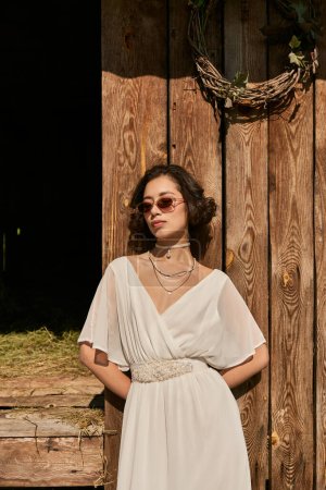 young asian bride in white wedding dress and sunglasses standing near wooden barn in countryside