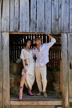 recién casados en el campo, novia asiática en botas de vaquero y vestido blanco de pie con el novio en el granero