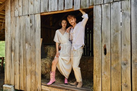 Photo for Stylish newlyweds, pretty asian bride in cowboy boots and white dress standing with groom in barn - Royalty Free Image