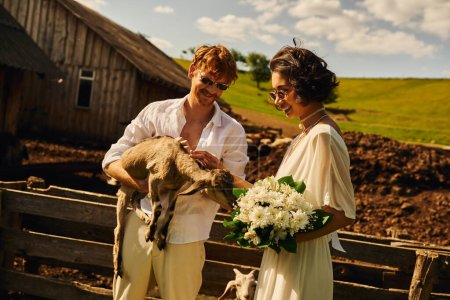 glückliches multiethnisches Brautpaar kuschelt süße Baby-Ziege, asiatische Frau in Hochzeitskleid und Sonnenbrille