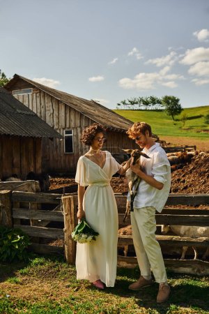 happy multiethnic couple in wedding gown and sunglasses cuddling cute baby goat, countryside