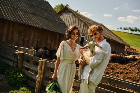 Lächelndes multiethnisches Paar in Brautkleid und Sonnenbrille, das niedliche Ziegenbaby kuschelt, Landschaft