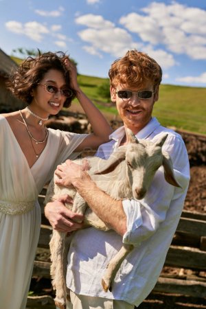 rustykalny ślub w stylu boho, szczęśliwy mężczyzna gospodarstwa dziecko kozy w pobliżu młoda azjatycka kobieta w sukni ślubnej