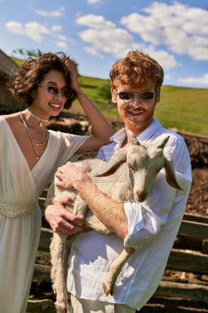 Foto de Boda rústica en estilo boho, hombre feliz sosteniendo bebé cabra cerca de mujer asiática joven en vestido de novia - Imagen libre de derechos