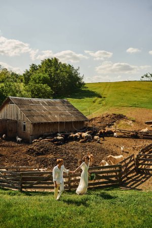 rustic wedding in boho style, happy groom holding hands with asian bride and walking near livestock