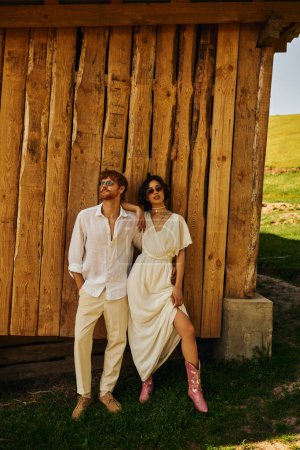 rustic wedding in boho style, stylish asian bride in wedding dress standing with groom in sunglasses