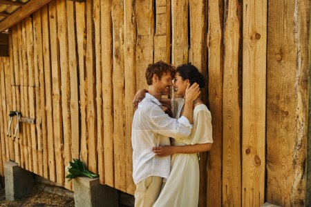 Boho-Hochzeit, glücklicher Bräutigam flirtet mit asiatischer Braut im weißen Kleid, Brautpaar auf dem Land