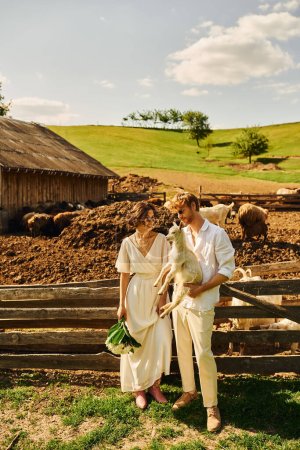 Téléchargez les photos : Mariage de style boho dans la campagne, marié heureux tenant bébé chèvre près de mariée asiatique en robe blanche - en image libre de droit