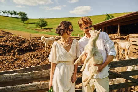 Photo for Just married couple, redhead groom holding baby goat near asian bride in white dress, boho style - Royalty Free Image