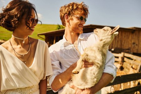 just married couple, redhead groom cuddling baby goat near asian bride in white dress, boho style