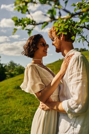 Foto de Estilo boho, felices recién casados interracial en gafas de sol y vestido de novia abrazándose en el jardín verde - Imagen libre de derechos