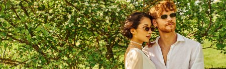 Téléchargez les photos : Jeunes mariés multiethniques en lunettes de soleil et robe de mariée étreignant dans le jardin vert, bannière - en image libre de droit