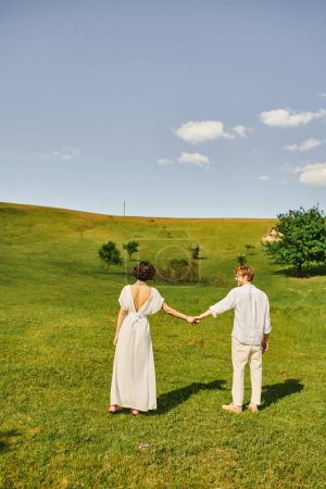 happy redhead man holding hands with bride in white dress and standing together in green field