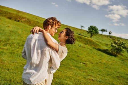 Téléchargez les photos : Heureux jeunes mariés souriant et étreignant dans le champ vert, mariée asiatique en robe de mariée et marié rousse - en image libre de droit