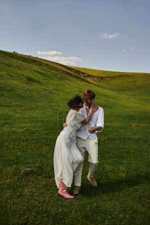 happy newlyweds, asian bride in white dress and cowboy boots walking with groom in green field