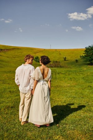 just married couple, asian bride in wedding dress holding hands with redhead groom in green field