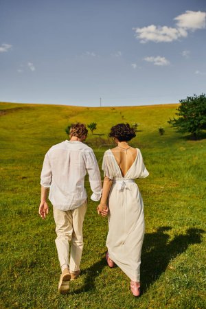 just married couple, back view of bride in wedding dress walking with groom in green field
