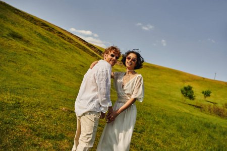 Foto de Boda rural en el campo, multiétnicos recién casados en vestido de novia mirando a la cámara en el campo verde - Imagen libre de derechos