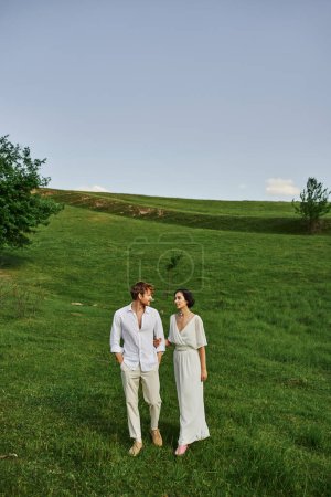 scenic landscape, young newlyweds in wedding gown walking together in green field, just married