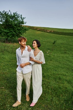 scenic landscape, happy newlyweds in wedding gown walking together in green field, just married