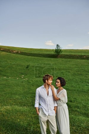 just married multiethnic couple standing together in green field, scenic and tranquil landscape