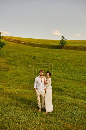 just married multicultural couple standing together in green field, scenic and tranquil landscape