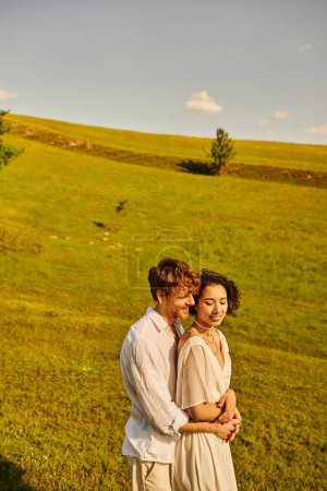 Téléchargez les photos : Couple juste marié, homme heureux embrassant mariée asiatique dans le champ vert, paysage pittoresque et tranquille - en image libre de droit