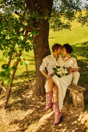 rothaariger Bräutigam umarmt asiatische Braut mit Strauß, während er auf Bank unter Baum sitzt, rustikale Hochzeit