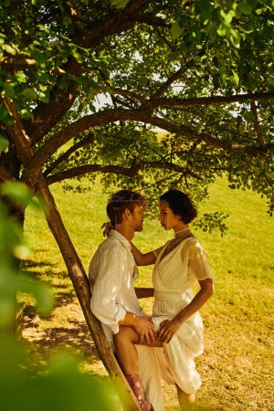 Foto de Vista lateral de pareja de estilo boho interracial cerca de escalera y árbol en el campo, boda rural - Imagen libre de derechos
