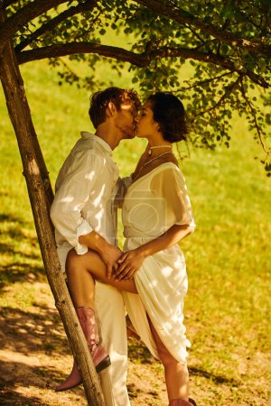 Foto de Interracial recién casados en estilo boho traje de novia besos bajo el árbol cerca de escalera, entorno rural - Imagen libre de derechos
