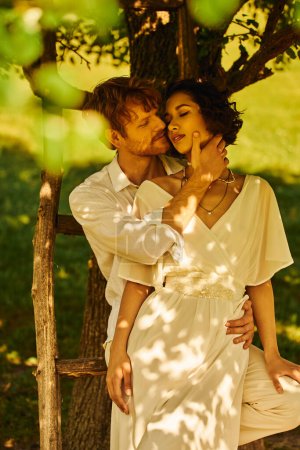 Photo for Happy redhead groom embracing tender asian woman in wedding dress under tree outdoors, rural setting - Royalty Free Image