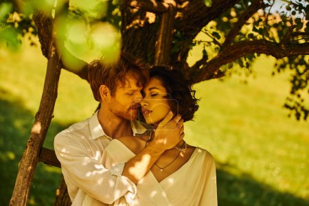 young redhead groom embracing asian bride in wedding dress under tree, countryside celebration
