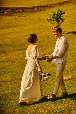 pelirroja novio y asiático novia con flores usando elegante traje boho y cogido de la mano en el campo