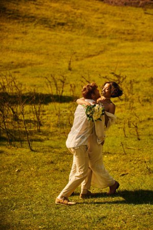 redhead groom embracing cheerful asian bride with flowers in green field, rural celebration