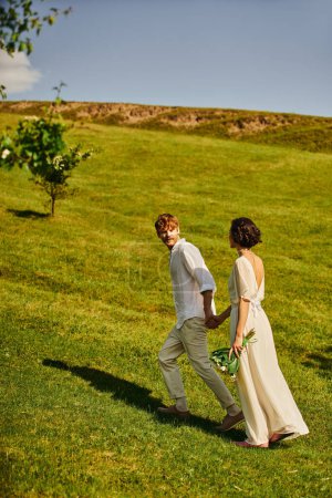 just married interracial couple in white wedding attire holding hands and running on green meadow