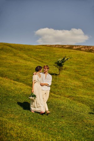 happy interracial newlyweds in boho style wedding attire walking in green field, scenic landscape