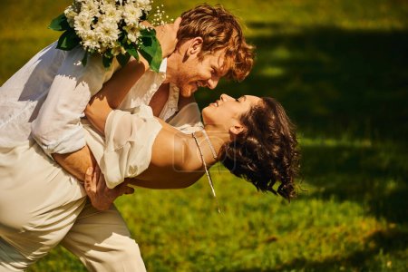 Téléchargez les photos : Vue latérale de rousse homme embrassant élégante mariée asiatique avec bouquet sur prairie verte, mariage rural - en image libre de droit