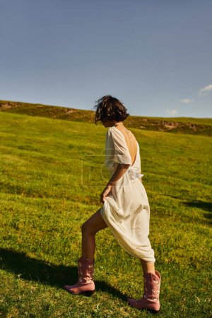 Photo for Brunette woman in white dress and cowboy boots walking under blue sky in green field, rural wedding - Royalty Free Image