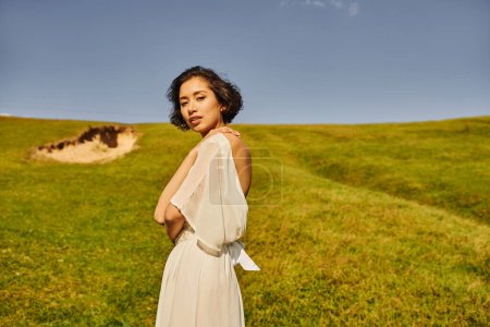 Téléchargez les photos : Brunette asiatique femme dans boho style robe de mariée en regardant la caméra dans le champ vert dans la campagne - en image libre de droit