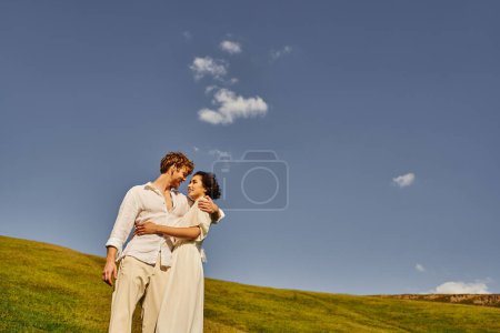 interracial newlyweds in boho style attire embracing under blue sky in green field, rustic wedding