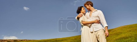 felices recién casados multiétnicos abrazándose en el prado verde bajo el cielo azul, boda en un entorno rural, pancarta