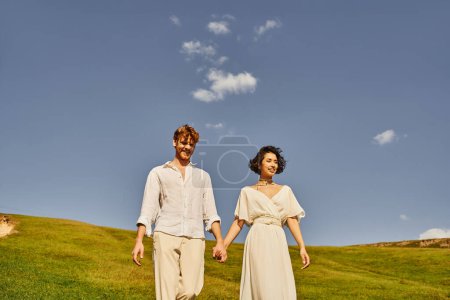 boda rústica, alegre pareja multiétnica en traje de estilo boho tomados de la mano y caminando en el prado