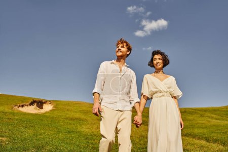 Foto de Jóvenes, despreocupados recién casados interracial tomados de la mano en el campo verde bajo el cielo azul, paisaje escénico - Imagen libre de derechos
