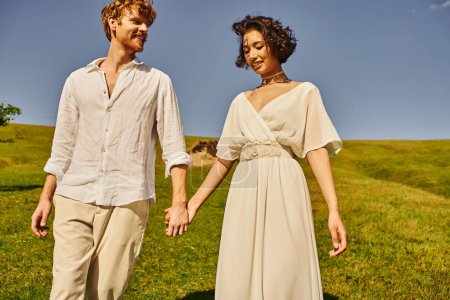 Téléchargez les photos : Moment heureux de jeunes mariés interracial en tenue de style boho tenant la main dans le champ, mariage rural - en image libre de droit