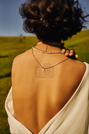 youthful woman in necklaces and bridal gown with naked back in countryside, serenity and happiness